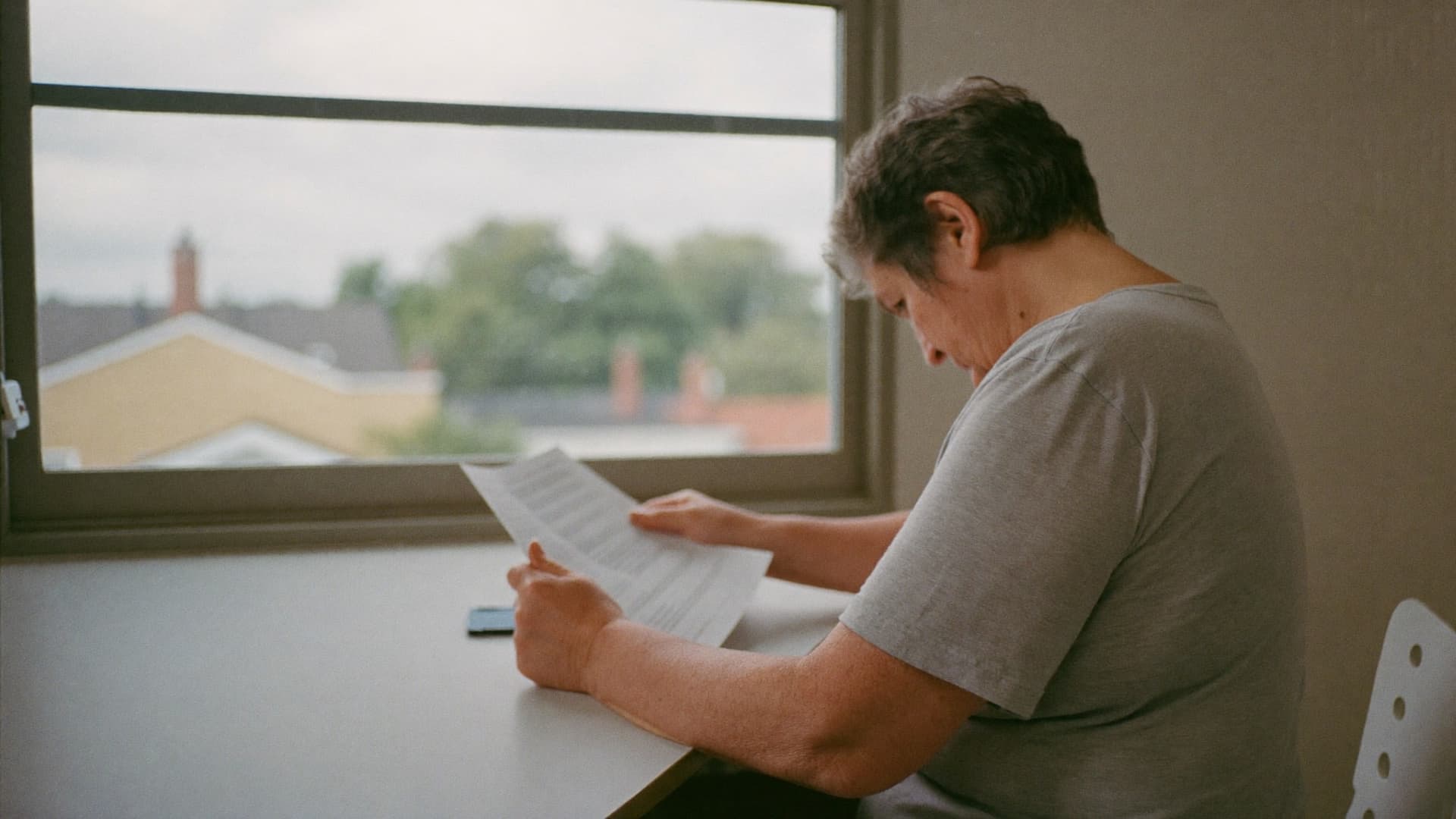 A woman reading a printed document at a desk beside a window.