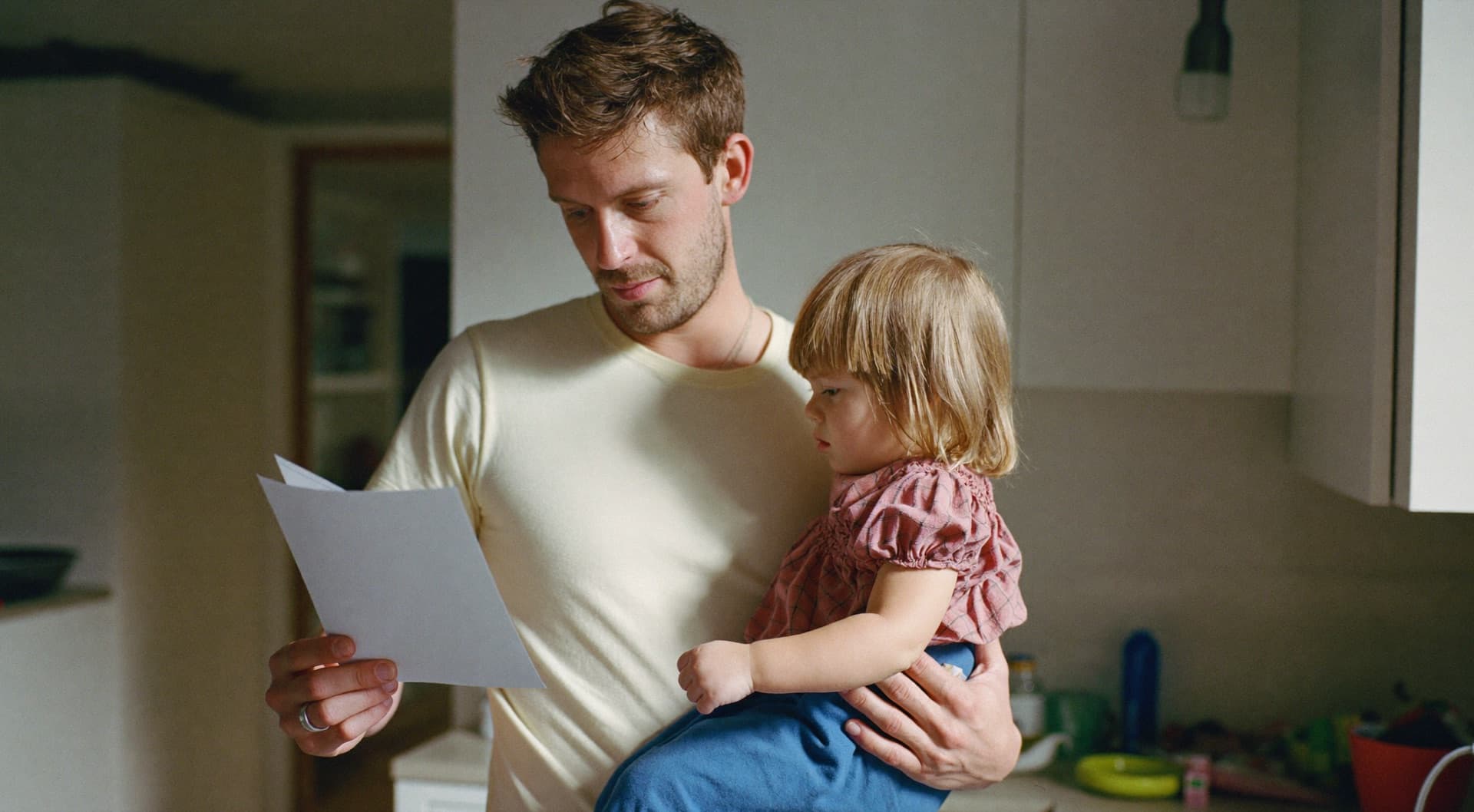A parent holding a toddler while reading a letter in a kitchen.
