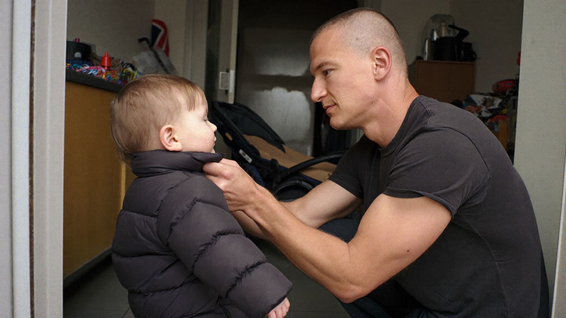 A parent crouching to fasten a small child's coat in a hallway, a pushchair behind them.